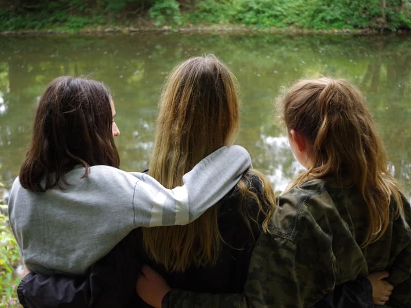 Drei Mädchen sitzen von hinten Arm in Arm am Ufer der Enz