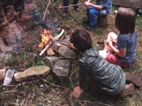 Drei Kinder sitzen von hinten um ein kleines Lagerfeuer zwischen Steinen